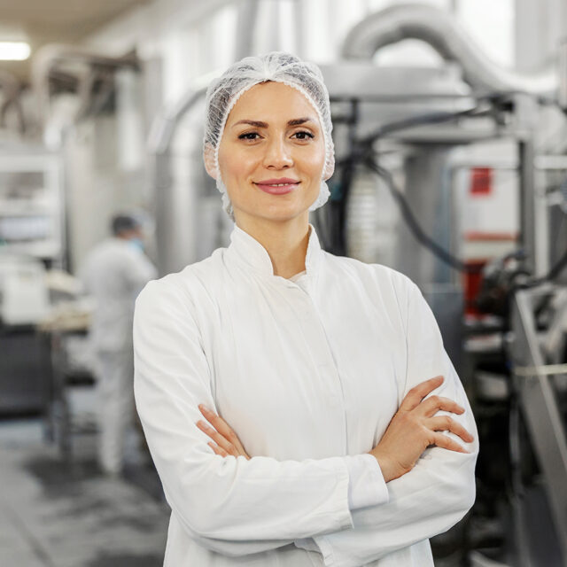 Lächelnde Frau mit Laborkittel und Haarnetz steht mit verschränkten Armen in Lebensmittelbetrieb