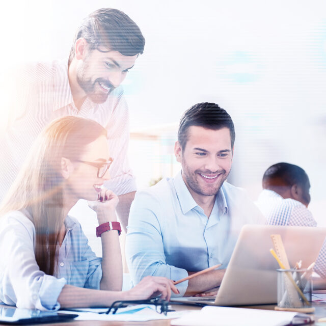 Teamarbeit: Bärtiger Mann steht über zwei jungen Mitarbeiter:innen, die in einem hellen Büro mit einem Laptop arbeiten.