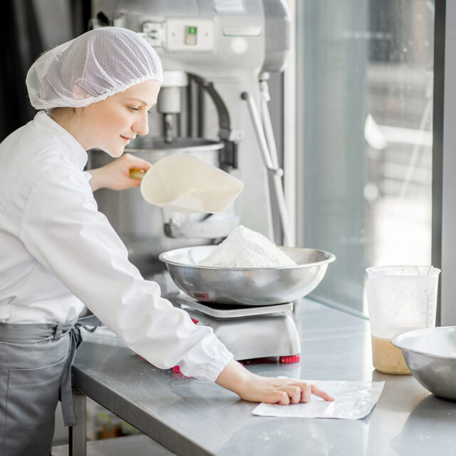 Konditorin in Arbeitskleidung wiegt Zutaten für Backwaren ab und arbeitet in der Backwarenherstellung.