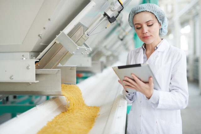Porträt einer jungen Frau von der Taille aufwärts, die neben Maschinen steht und mit einem digitalen Tablet die Makkaroni-Produktion in einer Lebensmittelfabrik überprüft