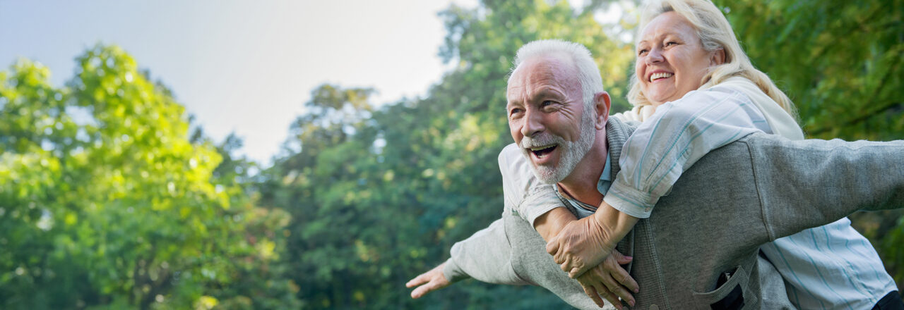 Zwei ältere Personen in grüner Natur; der Mann trägt die Frau huckepack, beide wirken aktiv und fröhlich.