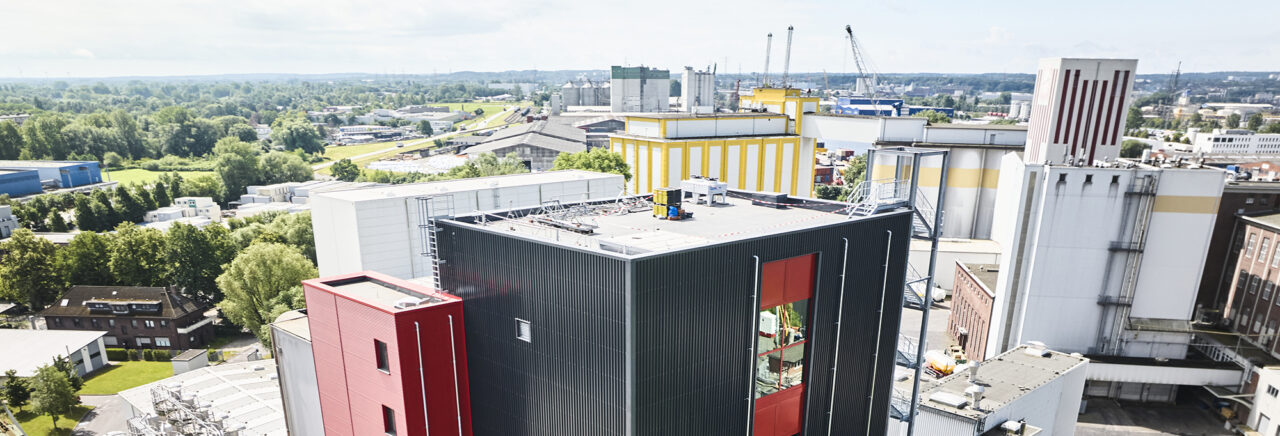 Industrieanlage mit schwarzen und roten Gebäuden im Vordergrund, gelben und weißen Hallen im Hintergrund sowie Kränen; umgeben von Grünflächen und kleineren Bauten