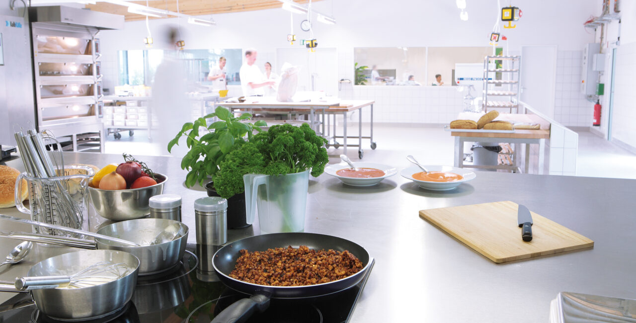 Blick in eine Industrieküche mit Kochutensilien, frischen Zutaten und mehreren Personen bei der Zubereitung von Speisen.
