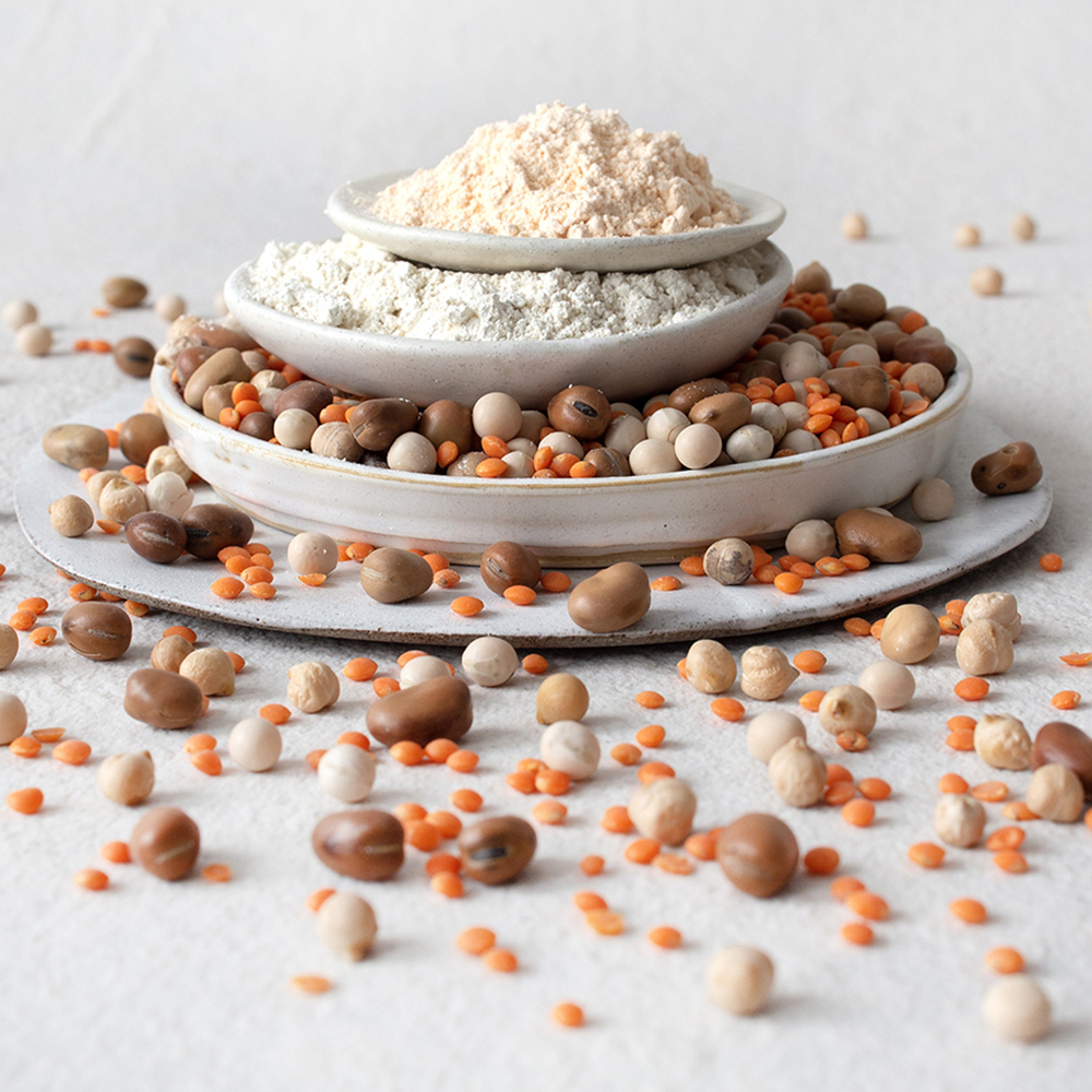 Drei gestapelte Teller mit Hülsenfruchtmehl, getrockneten Bohnen und Linsen; weitere Hülsenfrüchte liegen verstreut auf dem Untergrund.