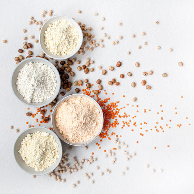 Vier Schalen mit Hülsenfruchtmehlen, weitere Hülsenfrüchte liegen verstreut auf dem Untergrund