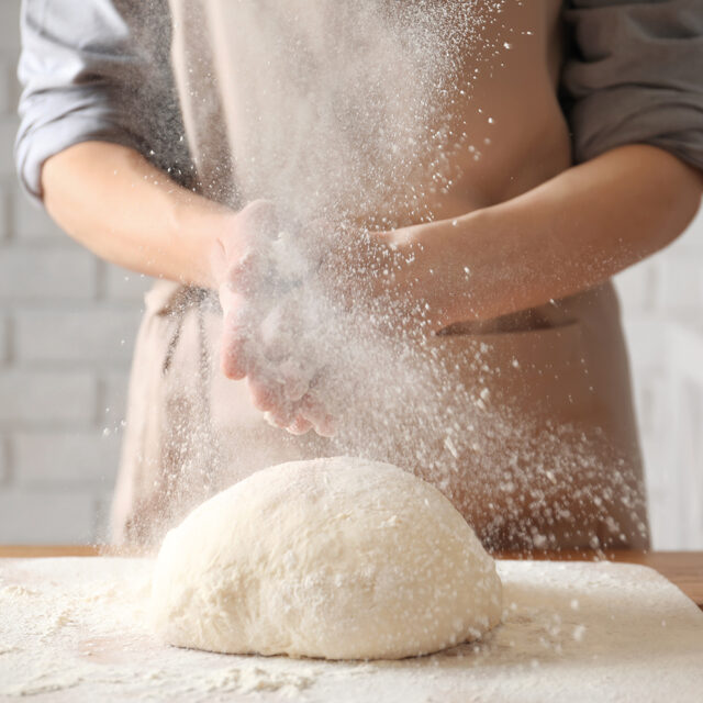 Frau klatscht in die Hände und streut Mehl über einen Teig auf einem Tisch